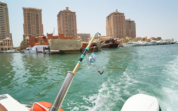 A fishing rod is mounted on a boat in the foreground during a Private Speed Boat Fishing Trip, with tall buildings, yachts, and covered boats along the waterfront in the background under a clear sky. The boat is moving, creating ripples in the water.