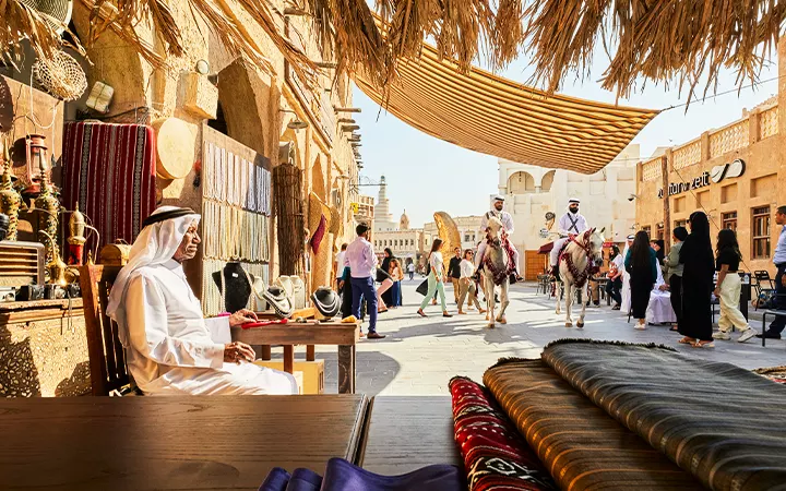A bustling Middle Eastern market scene with fabrics, traditional items, and people walking; a man in white robes sits at a stall in Souq Waqif, while two men on camels pass by under a fabric canopy near the Msheireb Museum Qatar.
