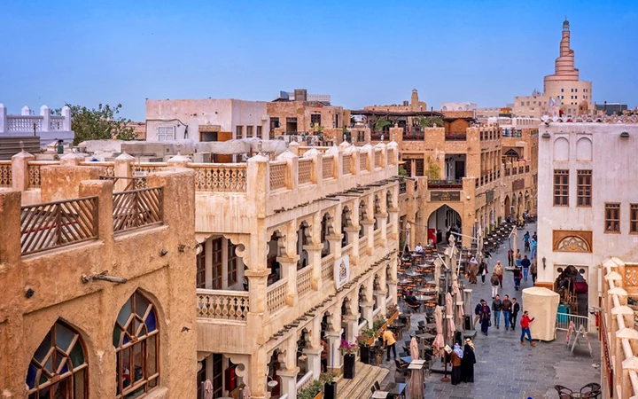A bustling street in Souq Waqif, lined with traditional tan buildings, invites people to walk and dine outdoors. The spiral mosque and clear blue sky set the scene, just a short stroll from the insightful Msheireb Museum Qatar.