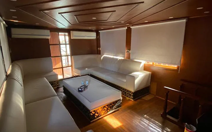 A cozy living room with wooden walls and ceiling, white leather sectional sofas, a central coffee table, two air conditioners, and large windows with blinds letting in sunlight—perfect for relaxing after a Traditional Dhow Boat Rental adventure.