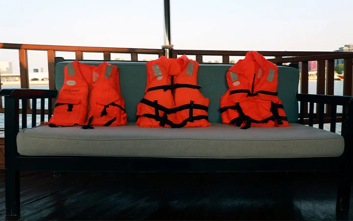 Three orange life jackets are placed side by side on a cushioned outdoor bench aboard a Traditional Dhow Boat Rental, with a wooden railing and water visible in the background.