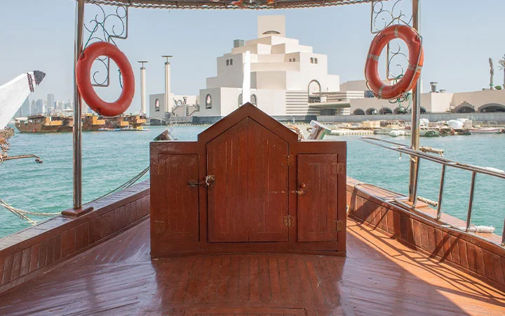 View from the deck of a traditional dhow boat rental with two orange life rings, looking toward the Museum of Islamic Art and city skyline across the water in Doha, Qatar, on a sunny day.