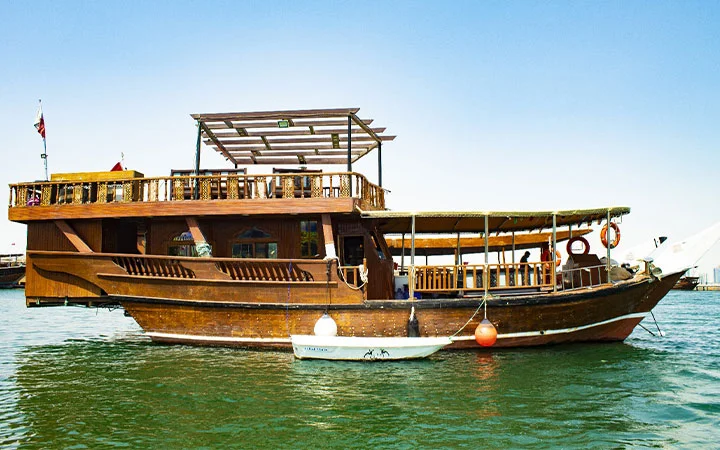 A traditional dhow boat rental with two levels floats on calm green water, a small white dinghy tied alongside. The boat features open seating areas, life rings, and flags, all beneath a clear blue sky.