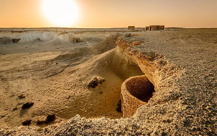 Desert landscape at sunset with ancient round stone structures, reminiscent of those near Zekreet Fort Qatar, partially buried in sand. A few rectangular buildings stand in the distance beneath a clear sky.