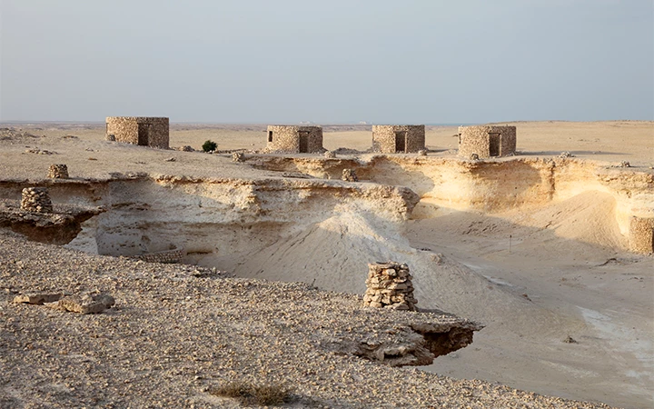 Stone huts stand on a barren, rocky desert landscape near the eroded cliffs of Zekreet Fort, Qatar, with pale sand under a hazy sky. The area appears remote and arid, with no vegetation visible nearby.