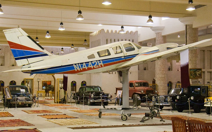A small white, blue, and red airplane is mounted indoors, surrounded by vintage cars and rugs in a spacious, well-lit museum—evoking the historic charm of places like Zekreet Fort Qatar.
