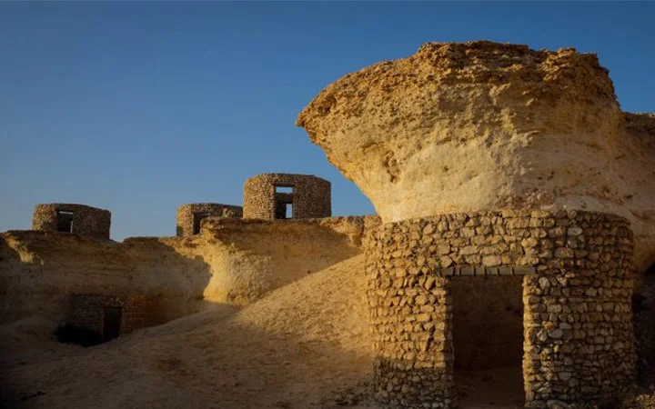 Ancient stone structures with round towers and weathered walls stand under a clear blue sky at Zekreet Fort, Qatar, partially built into a sandy, rocky hill. Sunlight casts strong shadows across the historic ruins.