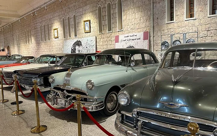 A row of vintage classic cars displayed in a museum, separated by red rope barriers; stone walls and vintage posters evoke the charm of sites like Zekreet Fort Qatar in the background.