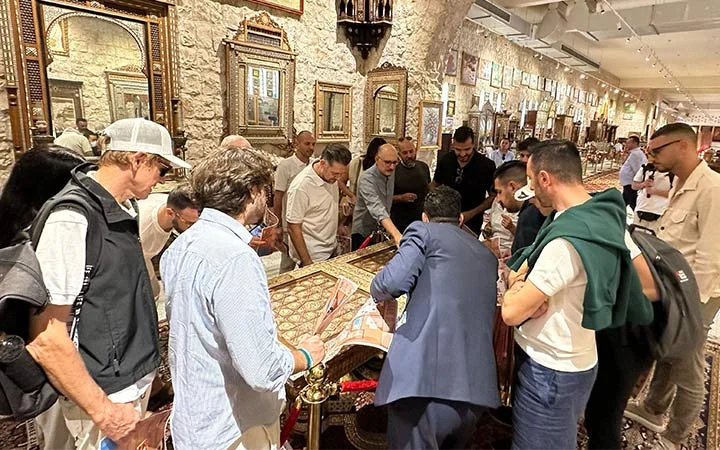 A group of people gathers around a display table inside a shop filled with ornate mirrors, framed art, and decorative items inspired by the designs of Zekreet Fort Qatar, as they examine items and chat with a salesperson.
