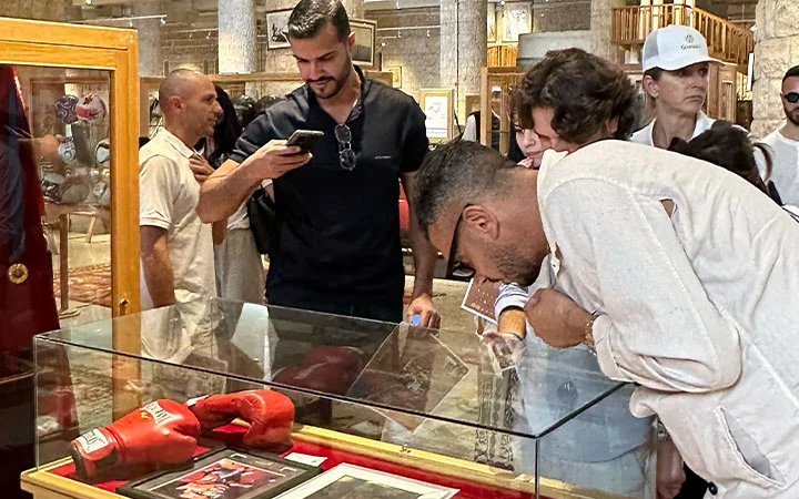 A group of people closely examine red boxing gloves and photographs displayed in a glass case at a museum or exhibition, reminiscent of the curious atmosphere found at Zekreet Fort Qatar. One man takes a photo while others lean in for a better look.