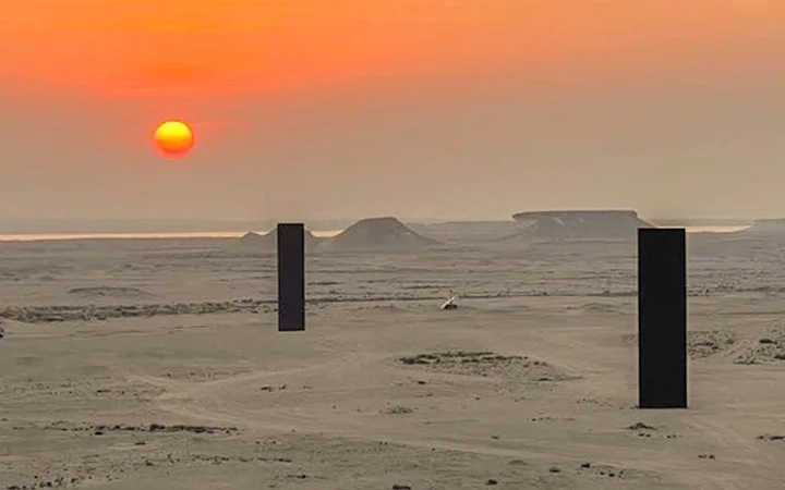 A desert landscape at sunset with a hazy orange sky, distant mesas, and three large, black rectangular monoliths standing upright on the sandy ground, reminiscent of the enigmatic scenery near Zekreet Fort in Qatar.