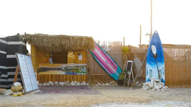 A small bamboo and straw surf shack with two colorful surfboards propped outside, a large white menu board, and a rustic fence, all set on a sandy beach near Purple Island Mangroves Kayaking.