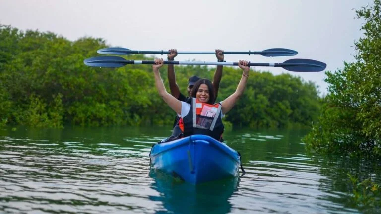 Two people in life jackets paddle a blue kayak through the calm, green waters of Purple Island Mangroves; lush greenery surrounds them as the woman in front smiles and raises her paddle above her head.