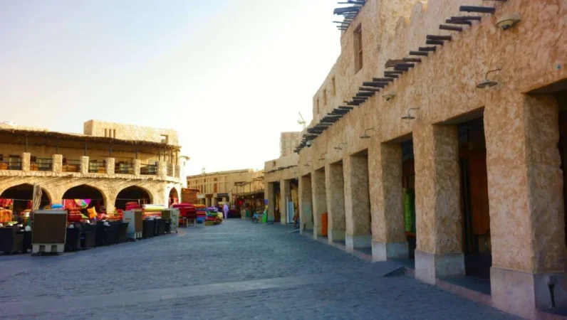 A sunlit street with traditional stone buildings and archways in a Middle Eastern market invites you to explore Doha City. Outdoor tables and chairs sit near shops, with colorful items visible in the distance along cobblestone walkways.