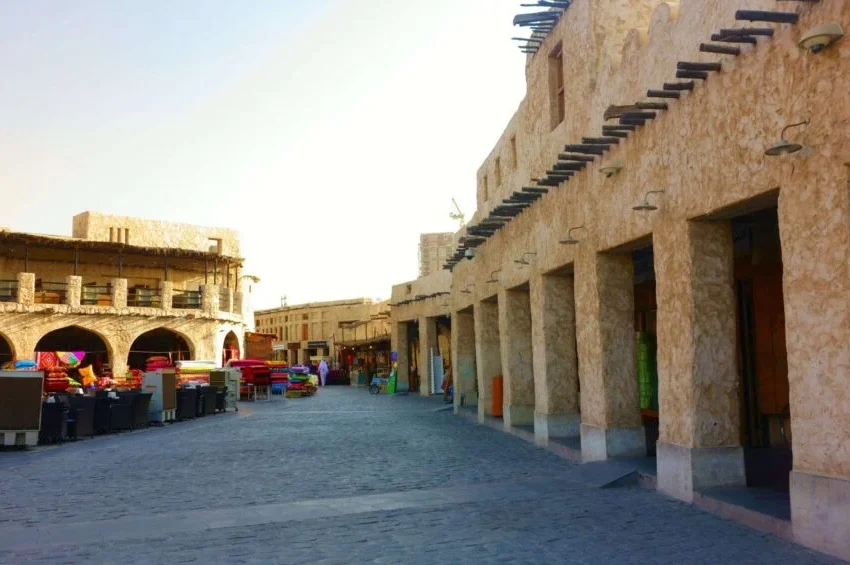 A sunlit street with traditional stone buildings and archways in a Middle Eastern market invites you to explore Doha City. Outdoor tables and chairs sit near shops, with colorful items visible in the distance along cobblestone walkways.