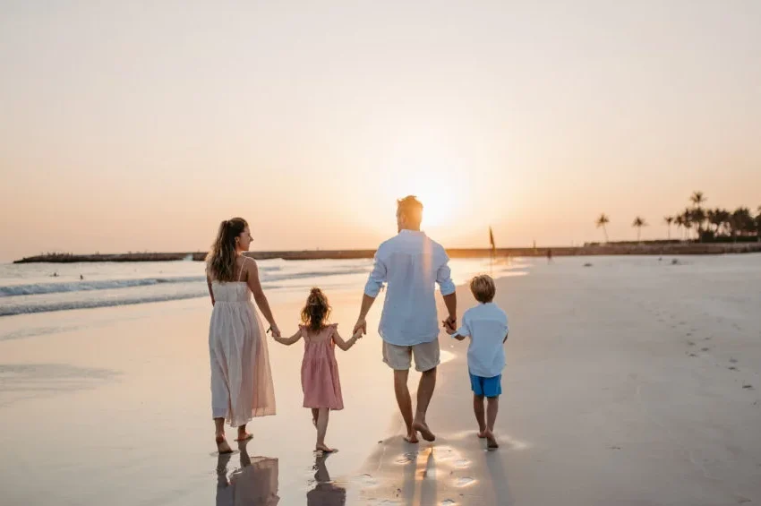 A family of four walks hand-in-hand along the beach at sunset, enjoying peace of mind knowing health insurance is mandatory, as the sun dips low on the horizon and palm trees sway gently in the distance.