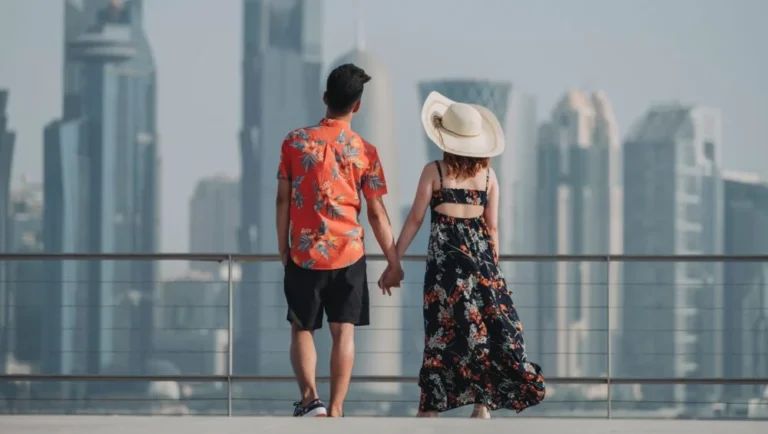 A couple holding hands stands on a terrace, facing a modern city skyline—perhaps dreaming of weddings in Qatar. The man wears a floral shirt and shorts, while the woman wears a floral dress and a wide-brimmed hat.