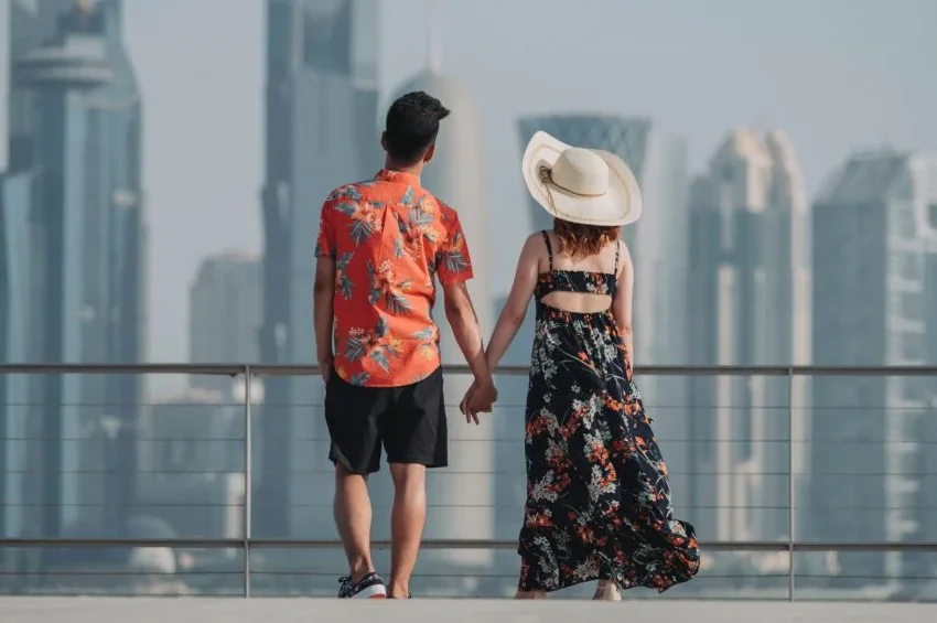 A couple holding hands stands on a terrace, facing a modern city skyline—perhaps dreaming of weddings in Qatar. The man wears a floral shirt and shorts, while the woman wears a floral dress and a wide-brimmed hat.