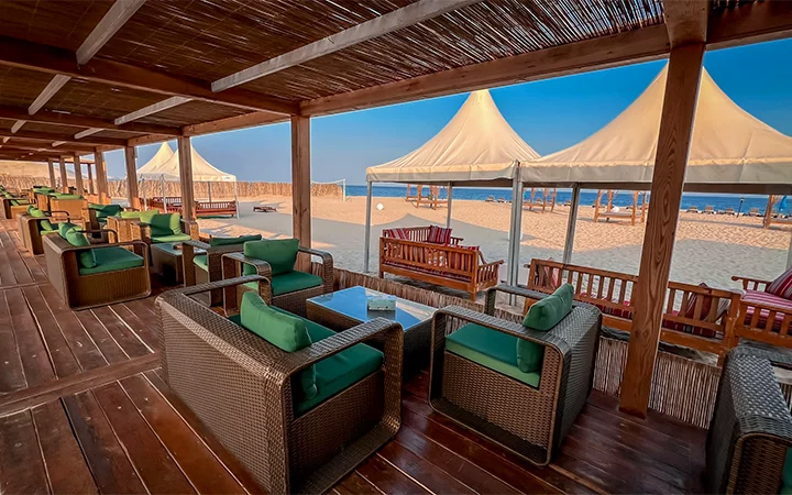 Outdoor lounge area with wicker chairs and green cushions on a wooden deck, overlooking a sandy beach—perfect for relaxing after a beachside adventure—where white canopy tents and a clear blue sky set the scene for an unforgettable extravaganza.