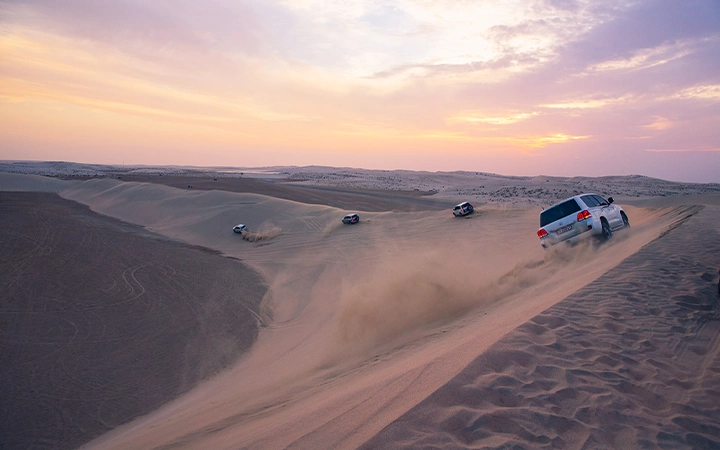 Several SUVs drive across sand dunes at sunset, kicking up dust as they climb and descend the sandy slopes, creating a true Desert Safari adventure under a colorful sky.