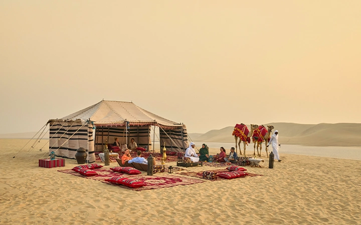 A group of people relax on colorful cushions outside a large tent during a Desert Safari adventure, with two camels nearby. The sky is hazy and sand dunes stretch into the background.