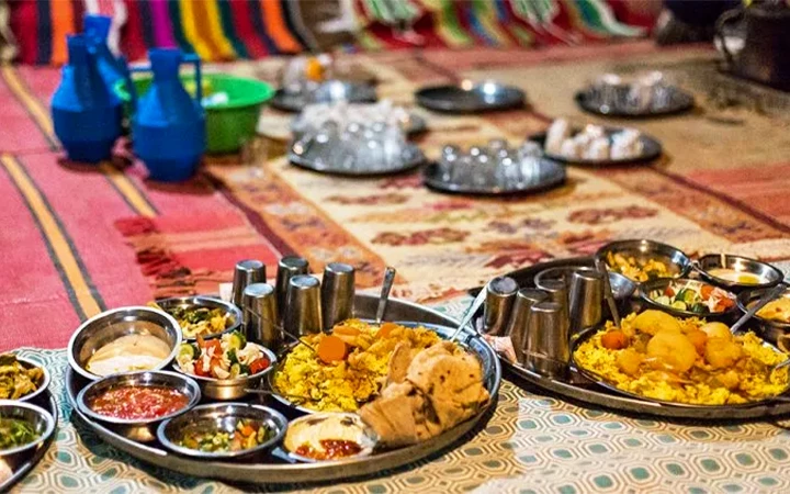 Two large round trays with various Indian dishes—curries, rice, bread, and sauces—are set on a colorful rug, creating a culinary extravaganza reminiscent of an adventure after a desert safari. Plates, glasses, and blue water jugs fill the background.