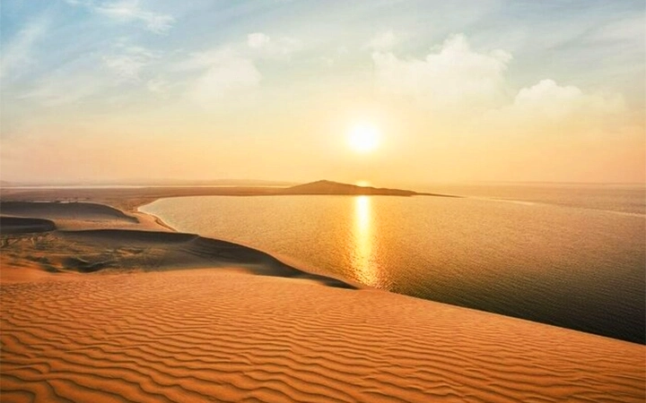 The sun sets over a calm body of water, casting a golden reflection. In the foreground are rippled sand dunes, evoking the thrill of a Desert Safari adventure, while distant hills line the horizon under a partly cloudy sky.