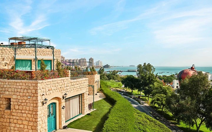A stone building with terraces overlooks a lush green lawn, trees, and a path, with the sea and modern city buildings in the background—an oasis of tranquility reminiscent of a half day desert safari Qatar adventure under a clear blue sky.