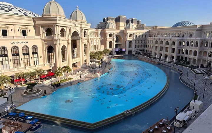 A large, elegant shopping complex with grand domes and arches features a central area with a decorative, curved water pool—perfect for relaxing after a half day desert safari Qatar—surrounded by outdoor seating, palm trees, and a Louis Vuitton store on the left.