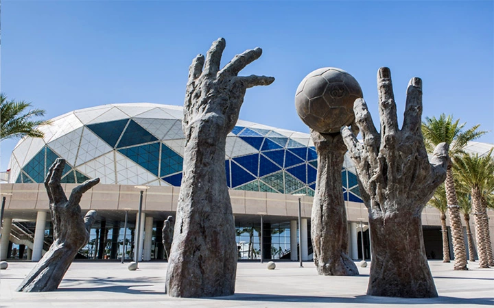 Large bronze sculptures of hands, one holding a soccer ball, stand outside a modern building with a geometric glass facade. Palm trees and clear blue sky evoke the adventurous spirit of a half day desert safari Qatar experience.