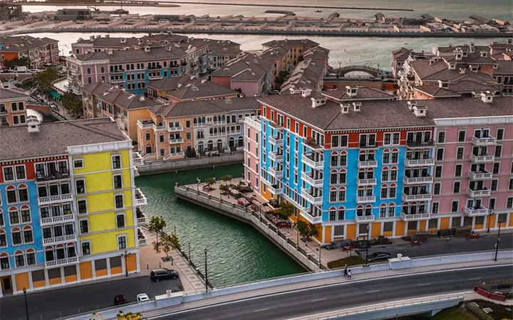 Colorful Mediterranean-style buildings line a canal with turquoise water, intersected by a road, while the sea shines in the background—a scene as striking as a half day desert safari Qatar adventure.