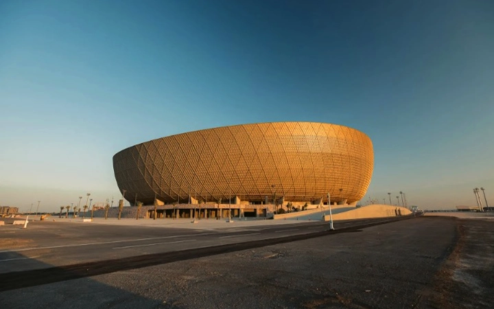 A large, circular stadium with a golden, lattice-like exterior facade gleams under a clear sky, bathed in warm sunlight—perfect before embarking on a half day desert safari Qatar adventure. An empty parking lot stretches out in the foreground.