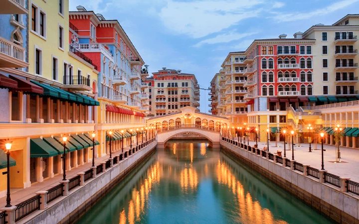 A canal lined with colorful buildings and street lamps reflects in the water, leading to an arched pedestrian bridge under a blue evening sky—an atmosphere as enchanting as a half day desert safari Qatar, with vibrant architecture and calm surroundings.