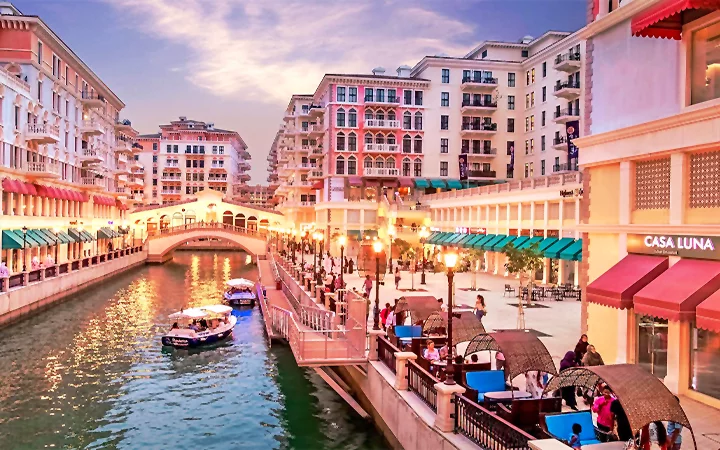 A vibrant canal scene at sunset, with boats on the water, people dining at outdoor cafes—much like the lively atmosphere after a half day desert safari Qatar adventurers might enjoy—colorful buildings and a pedestrian bridge complete the view.
