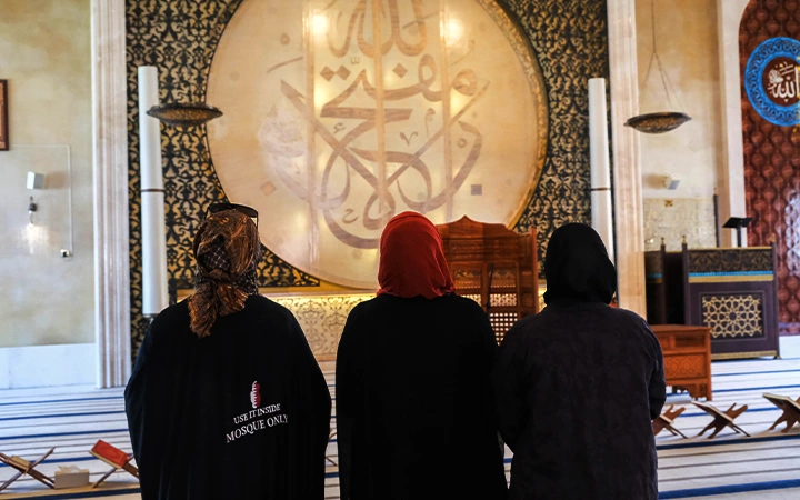 Three women with covered heads stand inside a mosque, facing a large decorative wall with Arabic calligraphy—a serene moment that contrasts with the thrill of a half day desert safari Qatar offers just beyond the city.