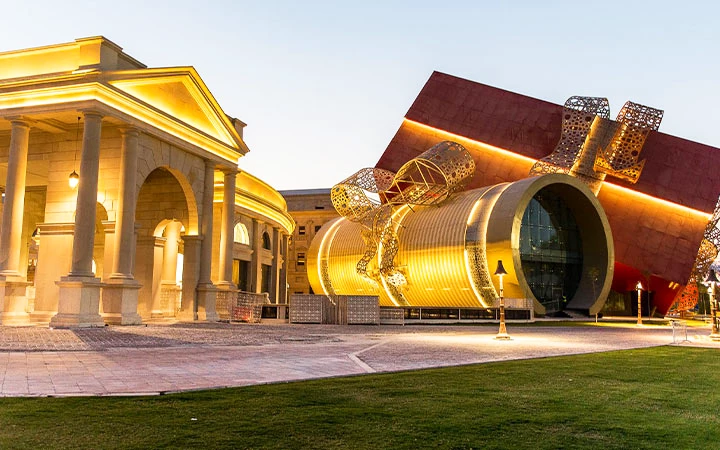A modern building shaped like a giant gift box with a bow, illuminated by golden lights, stands next to a classical structure with columns—reminiscent of the striking contrasts seen on a half day desert safari Qatar adventure.