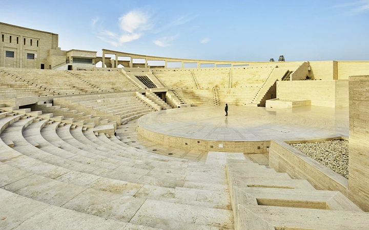 A person stands alone in the center of a large, open-air amphitheater made of light-colored stone, surrounded by tiered seating under a clear sky, reminiscent of moments found on a half day desert safari Qatar adventure.