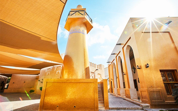 A tall, golden-yellow minaret rises among traditional Middle Eastern buildings under large shade sails, with bright sunlight and a clear blue sky—capturing the essence of Qatar before embarking on a half day desert safari adventure.