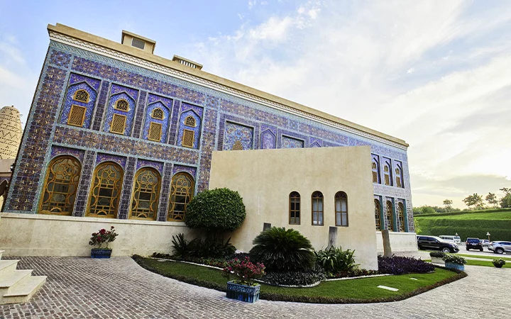 A large building with intricate blue and purple tilework, arched windows, and a beige extension stands near a manicured lawn and driveway—an inviting sight for those returning from a half day desert safari Qatar adventure.