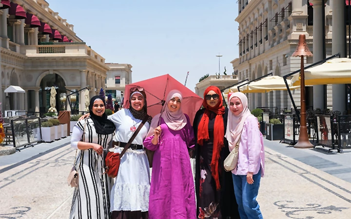 Five women in colorful, modest clothing and hijabs smile and pose together on a sunny street lined with elegant buildings and cafes, sharing stories of their recent half day desert safari Qatar. The atmosphere is cheerful and lively.