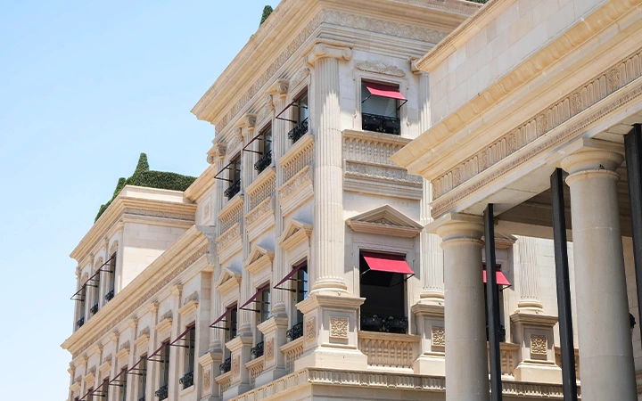A grand neoclassical building with tall columns, ornate stonework, and red-awninged windows stands proudly beneath a clear blue sky—an elegant contrast to the adventurous spirit of a half day desert safari Qatar offers.