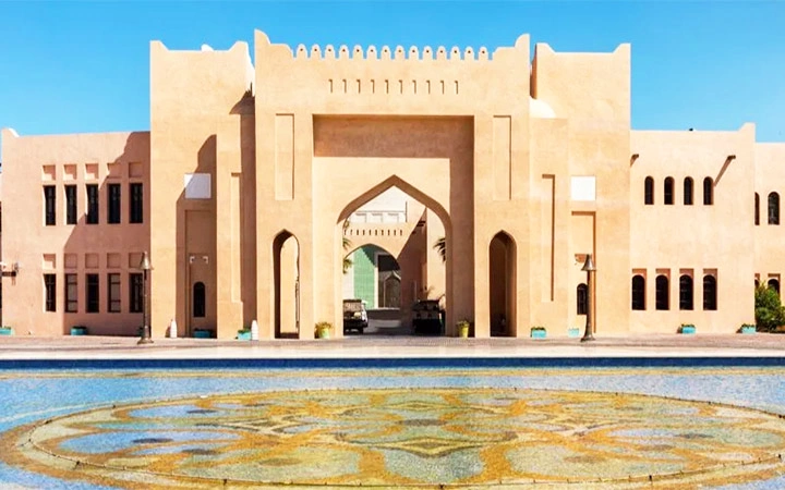 A traditional beige building with arched entrances and geometric patterns stands behind a decorative circular fountain, under a clear blue sky—an inviting scene before embarking on a half day desert safari Qatar adventure.