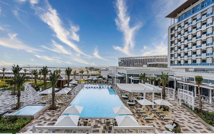 A modern hotel with a geometric facade overlooks a large outdoor pool—an inviting retreat for guests enjoying the Qatar Overnight Layover Package, surrounded by lounge chairs, umbrellas, and palm trees under a blue sky.