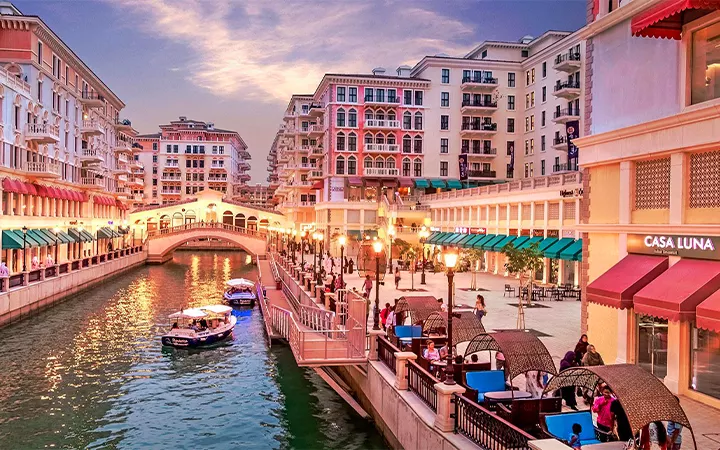 A picturesque canal lined with colorful buildings, outdoor cafes, and people enjoying a waterfront promenade at sunset—perfect for travelers seeking a relaxing stroll, much like moments found in a Qatar Overnight Layover Package.