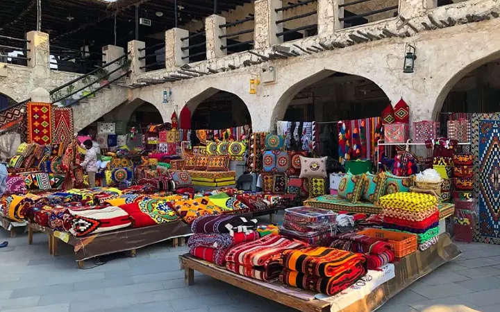 Colorful textiles, carpets, and cushions with intricate patterns are displayed on tables in front of a stone building with arches at an outdoor market—a vibrant scene you might explore during a Qatar Overnight Layover Package. Two people browse among the items.