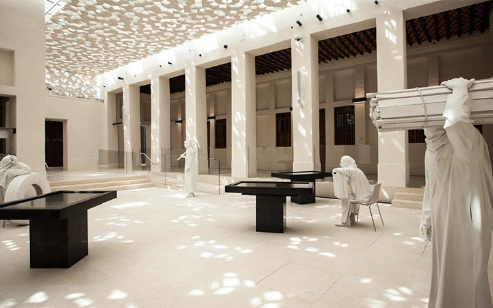 A bright, modern atrium—perfect for a Qatar Overnight Layover Package—features sculpted white figures performing work activities beneath sunlight filtering through a patterned ceiling and geometric shadows. Black tables are scattered throughout the space.