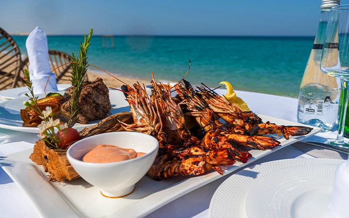 A platter of grilled seafood, including shrimp and prawns, is served with sauce and lemon on a white tablecloth at an outdoor beachside dining table—a perfect meal for those enjoying a Qatar Overnight Layover Package with ocean views.