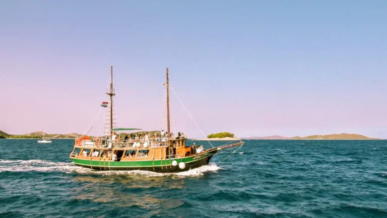 A wooden house boat in Qatar with passengers sails on blue-green water under a clear sky, small islands dotting the distance. A sailboat appears further back on the left side.