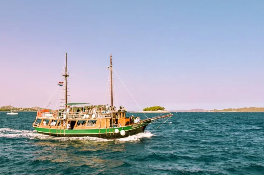 A wooden house boat in Qatar with passengers sails on blue-green water under a clear sky, small islands dotting the distance. A sailboat appears further back on the left side.