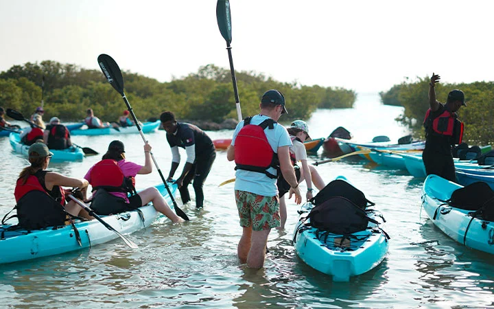 A group of people wearing life jackets prepare to kayak in shallow water, with several blue kayaks and lush green bushes in the background. Some are ready for their Full Moon Kayaking Qatar adventure, seated while others stand holding paddles.